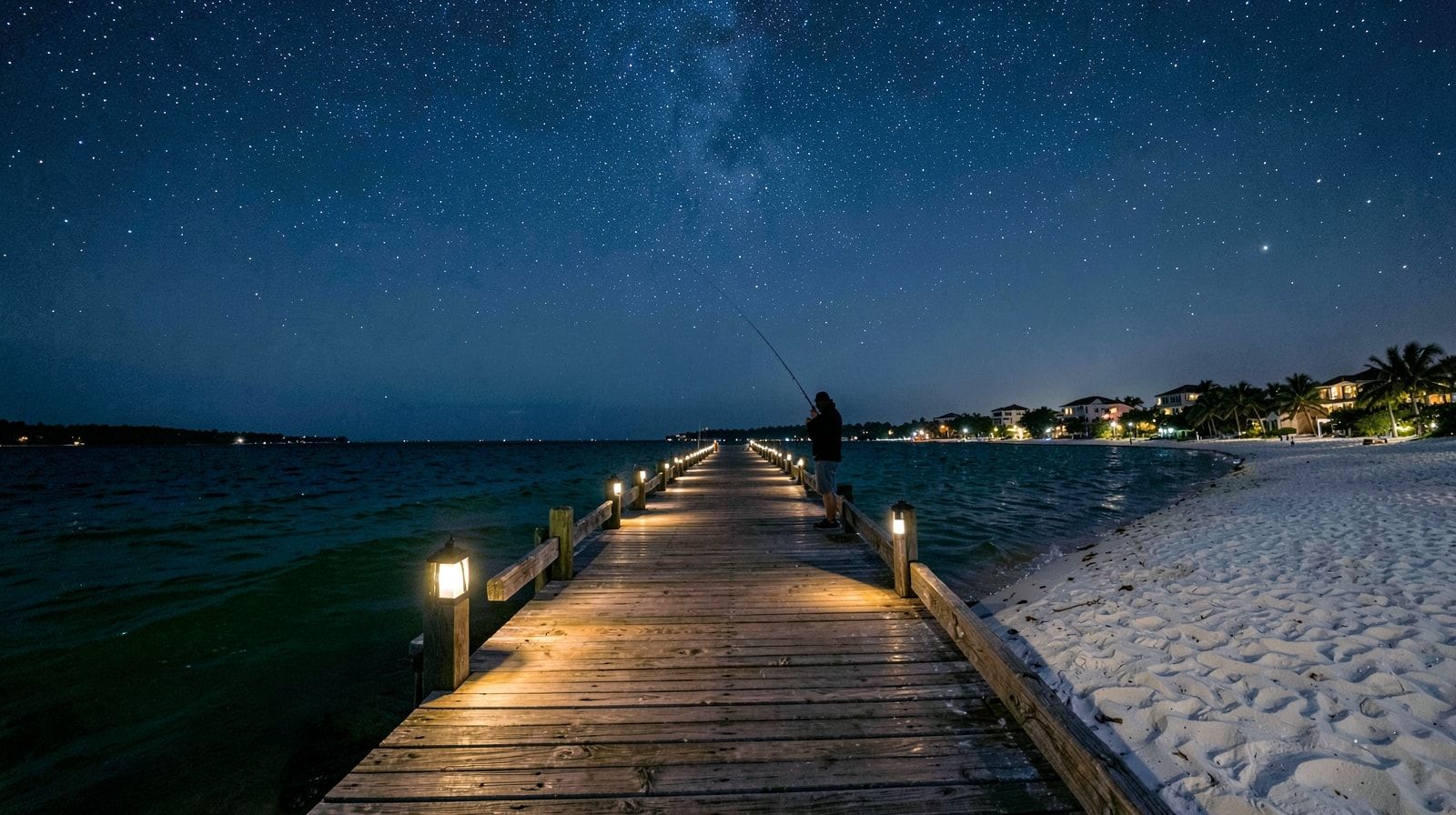 Solo angler fishing from a lit wooden pier over dark Gulf of Mexico water at night in the Florida Panhandle