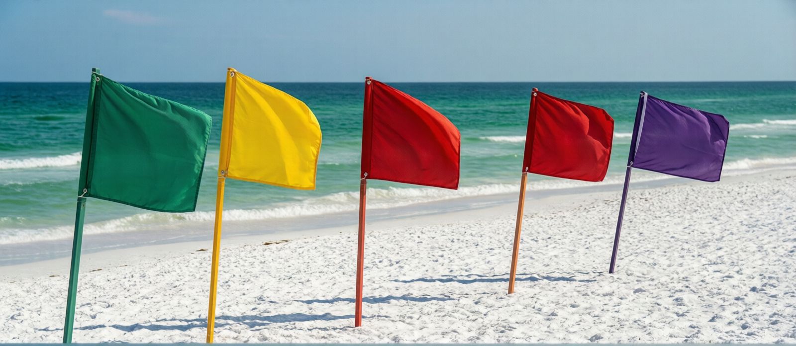 Five beach safety flags in green, yellow, single red, double red, and purple displayed on poles along a white sand Florida beach