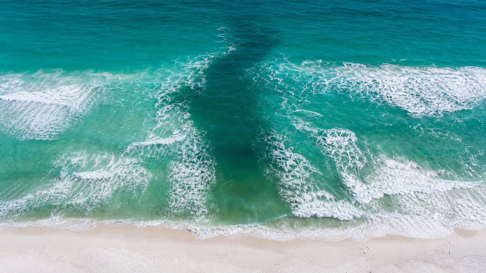 Aerial view of Destin beach showing a visible rip current as a dark calm channel cutting through the white breaking surf and extending seaward