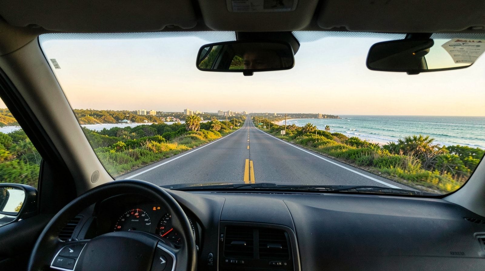View through a car windshield on a long road trip, straight interstate disappearing to the horizon through green summer countryside at dawn