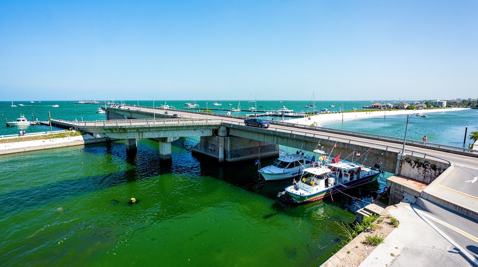 Car crossing a bridge over a coastal harbor with emerald green water and charter fishing boats below on a clear sunny day